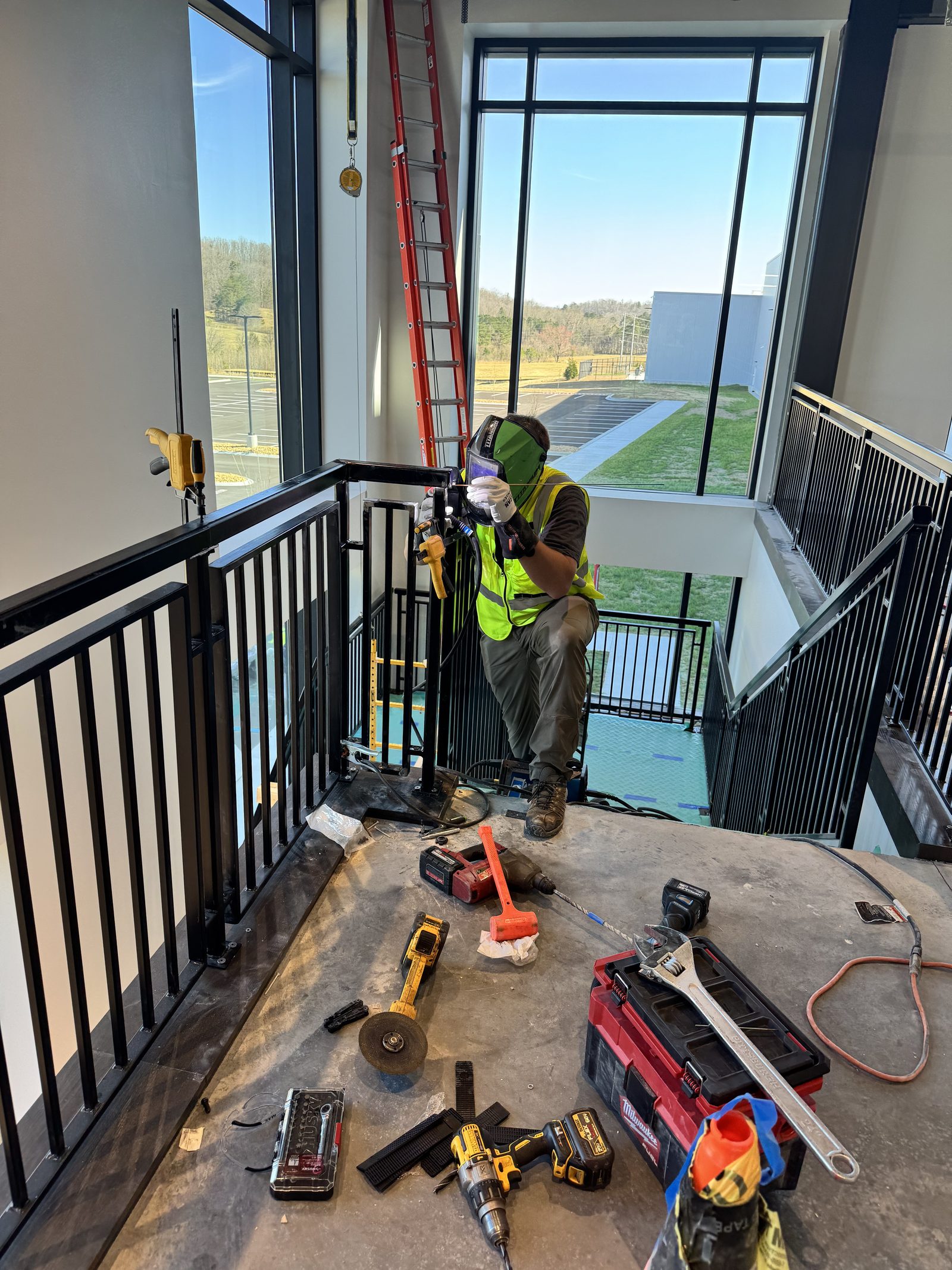Adam Smith in a welding hood and hi-vis vest, welding a steel stair rail in place on site, tools spread across the landing.
