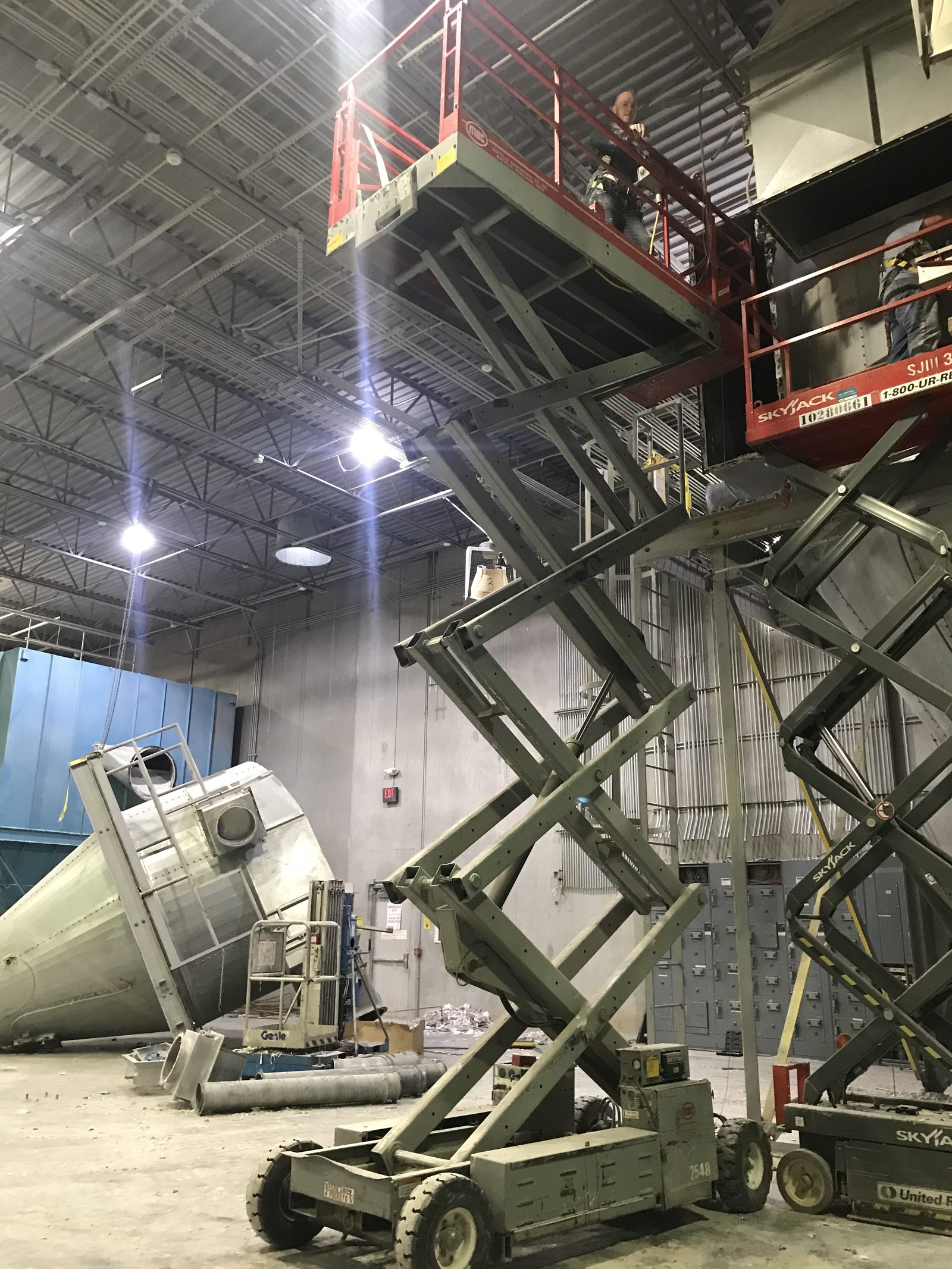 Two scissor lifts raised to the ceiling of an industrial plant during a tear-out, a large steel cyclone cut down and lying on the floor.